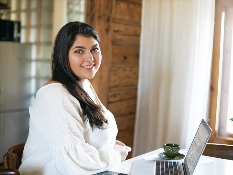 Side view of confident skillful young overweight female copywriter or translator sitting at table in front of open laptop, using wireless internet connection for distant work, smiling, drinking coffee